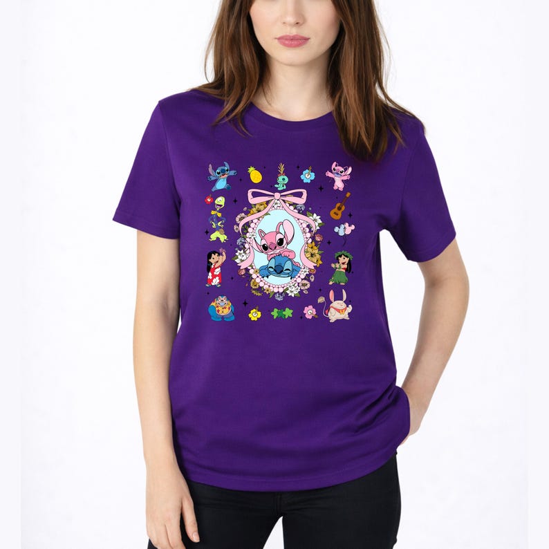 May include: A purple short-sleeved t-shirt featuring a central oval frame decorated with flowers and a pink ribbon, containing illustrations of two blue and pink alien characters. Smaller colorful character graphics surround the central frame.
