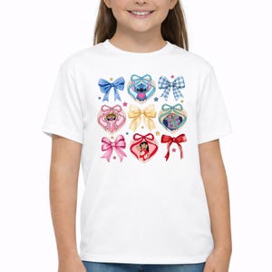May include: A white short-sleeved t-shirt featuring a central graphic print of various colorful bows and heart-shaped frames. Inside the hearts are illustrations of Stitch, Angel, and Lilo, accented by small stars scattered across the design.