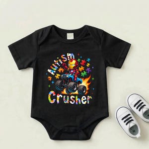 Puede incluir: Body de beb&eacute; negro con un gr&aacute;fico colorido de Iron Man conduciendo un cami&oacute;n monstruo, rodeado de piezas de rompecabezas y llamas. El texto dice "Autism Crusher". Zapatos de beb&eacute; blancos est&aacute;n al lado del body.