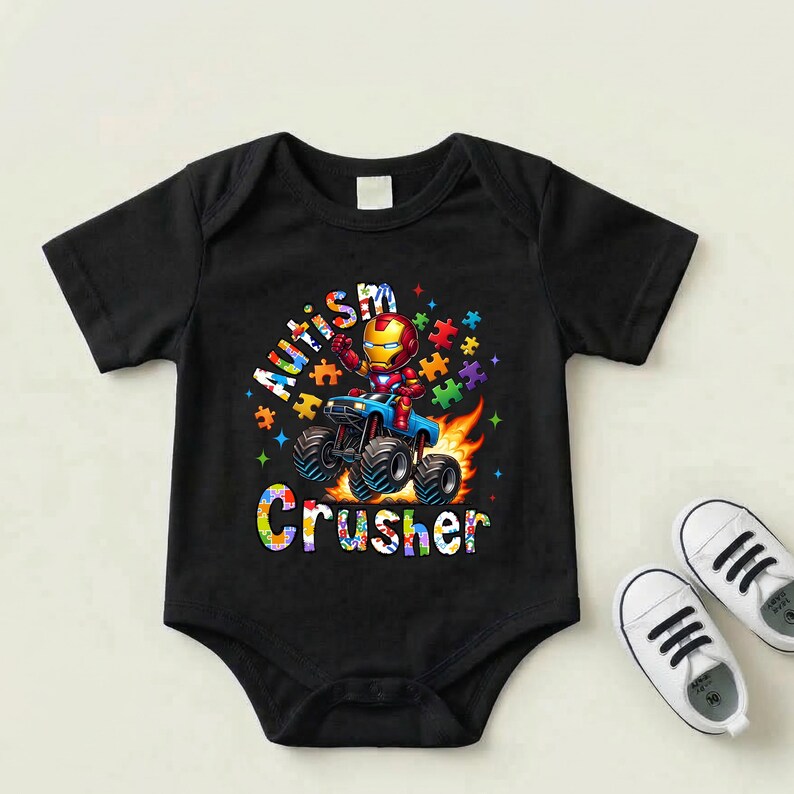 Puede incluir: Body de beb&eacute; negro con un gr&aacute;fico colorido de Iron Man conduciendo un cami&oacute;n monstruo, rodeado de piezas de rompecabezas y llamas. El texto dice "Autism Crusher". Zapatos de beb&eacute; blancos est&aacute;n al lado del body.