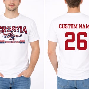May include: White t-shirt featuring "CROATIA" in red and white, with a soccer player graphic and "WORLD CUP 2026". The back displays "CUSTOM NAME" and the number "26" in red. A soccer-themed apparel item.