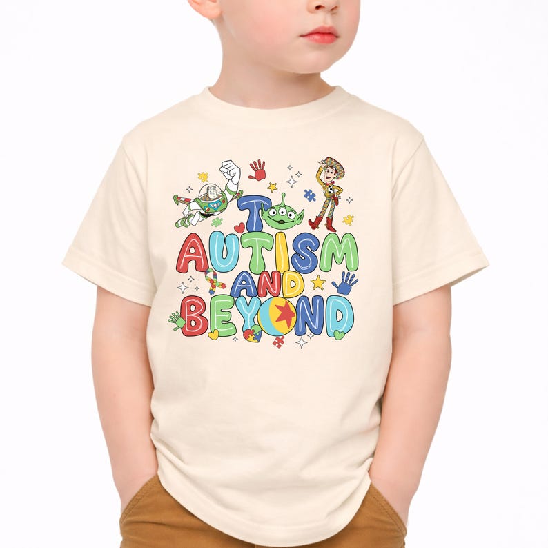 Op de afbeelding: Een cr&egrave;mekleurig T-shirt met de tekst "Autisme en verder" in kleurrijke, speelse letters. Het ontwerp bevat stripfiguren en handafdrukken. Het shirt wordt door een kind gedragen.