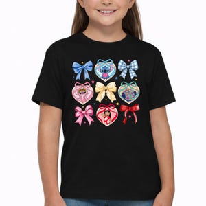 May include: A black short-sleeve t-shirt featuring a graphic print of various colorful bows and heart-shaped frames. Inside the hearts are illustrations of Stitch, Angel, and Lilo from Lilo and Stitch, accented by small scattered stars.