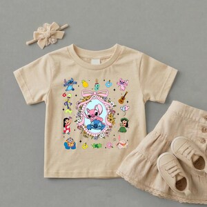 May include: A beige short-sleeved t-shirt featuring a central graphic of Stitch and Angel framed by a pink bow and floral wreath, surrounded by smaller character icons including Lilo, a guitar, and tropical elements, paired with a matching hair bow.