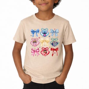 May include: A beige short-sleeve t-shirt featuring a graphic print of various bows and heart-shaped frames containing cartoon characters. The design includes blue, pink, and red bows, a central gold bow, and small colorful stars scattered throughout.