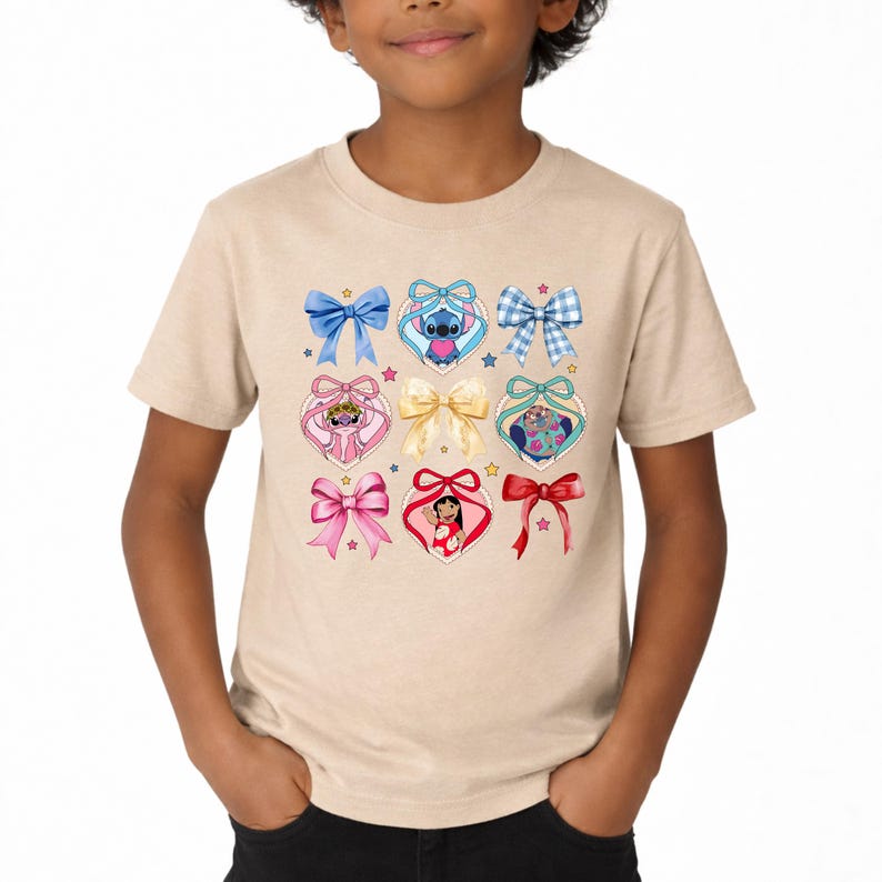 May include: A beige short-sleeve t-shirt featuring a graphic print of various bows and heart-shaped frames containing cartoon characters. The design includes blue, pink, and red bows, a central gold bow, and small colorful stars scattered throughout.