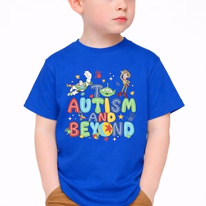 Op de afbeelding: Een koningsblauw t-shirt met de tekst "AUTISM AND BEYOND" in kleurrijke letters. Het shirt bevat cartoonfiguren en handafdrukken. Het t-shirt wordt door een kind gedragen.