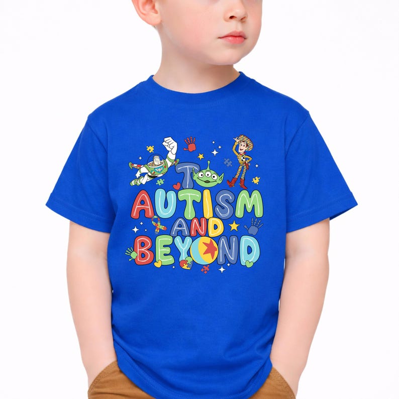 Op de afbeelding: Een koningsblauw t-shirt met de tekst "AUTISM AND BEYOND" in kleurrijke letters. Het shirt bevat cartoonfiguren en handafdrukken. Het t-shirt wordt door een kind gedragen.