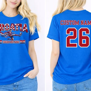 May include: Blue t-shirt with "CROATIA" in red and white, a soccer player graphic, and "WORLD CUP 2026". The back features "CUSTOM NAME" and the number "26" in red with a white outline.