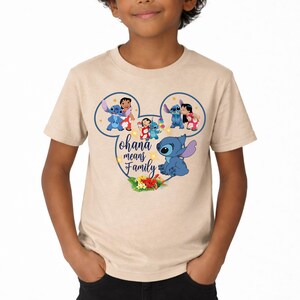 May include: A beige short-sleeve t-shirt featuring a graphic of Stitch and Lilo inside a Mickey Mouse ear outline. The design includes the text Ohana means Family, small yellow stars, and a cluster of tropical hibiscus flowers at the bottom.