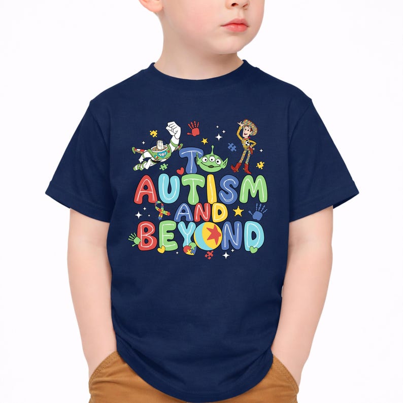 Op de afbeelding: Marineblauw T-shirt met de tekst "To Autism and Beyond" in kleurrijke, speelse letters. Het ontwerp bevat stripfiguren en symbolen die verband houden met autismebewustzijn. Een leuke en ondersteunende tee.