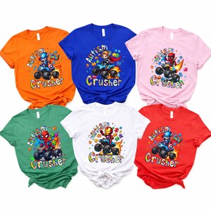 Puede incluir: Seis camisetas coloridas con gr&aacute;ficos de superh&eacute;roes. Cada camiseta presenta un superh&eacute;roe diferente conduciendo un monster truck con las palabras "Autism Crusher" en una fuente colorida. Las camisetas son naranja, azul, rosa, verde, blanca y roja.