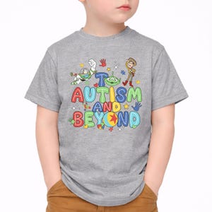 Op de afbeelding: Een grijs t-shirt met de tekst "Autisme en verder" in kleurrijke, speelse letters. Het ontwerp bevat stripfiguren en handafdrukken. Het t-shirt wordt door een kind gedragen.