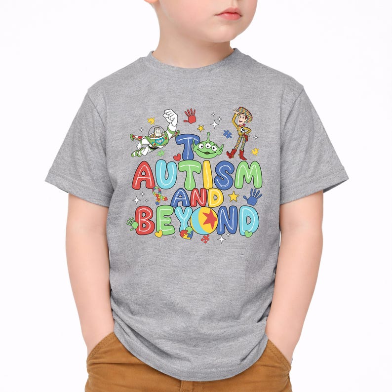 Op de afbeelding: Een grijs t-shirt met de tekst "Autisme en verder" in kleurrijke, speelse letters. Het ontwerp bevat stripfiguren en handafdrukken. Het t-shirt wordt door een kind gedragen.