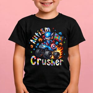 Puede incluir: Camiseta negra con un personaje de dibujos animados del Capit&aacute;n Am&eacute;rica conduciendo un cami&oacute;n monstruo azul. La camiseta tiene las palabras "Autism Crusher" y una bandera de rompecabezas. El dise&ntilde;o incluye llamas y piezas de rompecabezas coloridas.