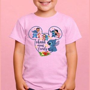 May include: A light pink short-sleeved t-shirt featuring a graphic of Stitch and Lilo inside a Mickey Mouse ear outline. The design includes the text Ohana means Family, small yellow stars, and a cluster of tropical flowers at the bottom.