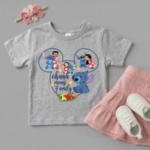 May include: A heather gray short-sleeve t-shirt featuring a graphic of Stitch and Lilo inside a Mickey Mouse ear outline. The text reads Ohana means Family, accompanied by colorful tropical flowers. A pink hair bow and a pink skirt are also shown.
