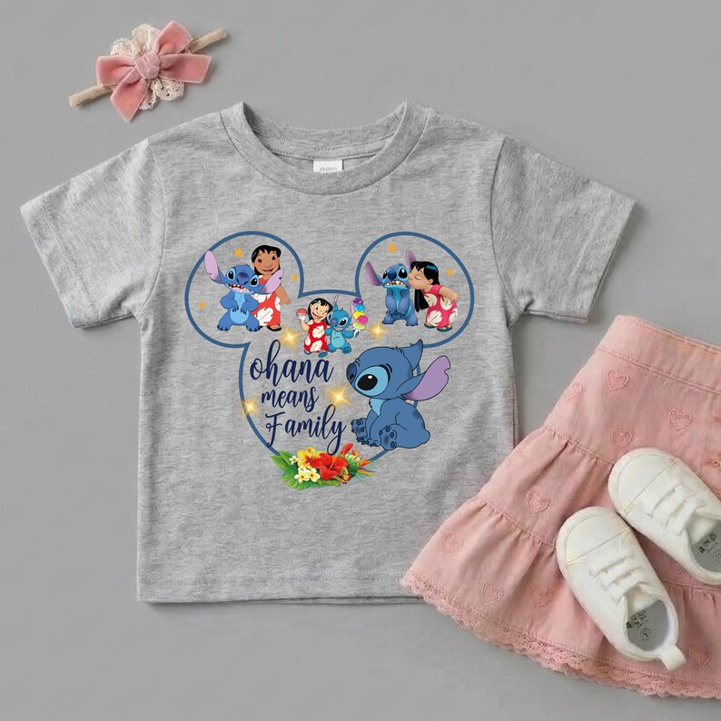 May include: A heather gray short-sleeve t-shirt featuring a graphic of Stitch and Lilo inside a Mickey Mouse ear outline. The text reads Ohana means Family, accompanied by colorful tropical flowers. A pink hair bow and a pink skirt are also shown.