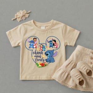 May include: A beige short-sleeved t-shirt featuring a graphic of Stitch and Lilo inside a Mickey Mouse ear outline with the text Ohana means Family, accompanied by a matching beige hair bow and a beige tiered skirt with two beige sneakers.