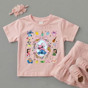 May include: A light pink short-sleeved t-shirt featuring a central oval frame with a pink bow, containing two blue and pink alien characters. The shirt is decorated with various small cartoon figures, flowers, a guitar, and a pineapple graphic.