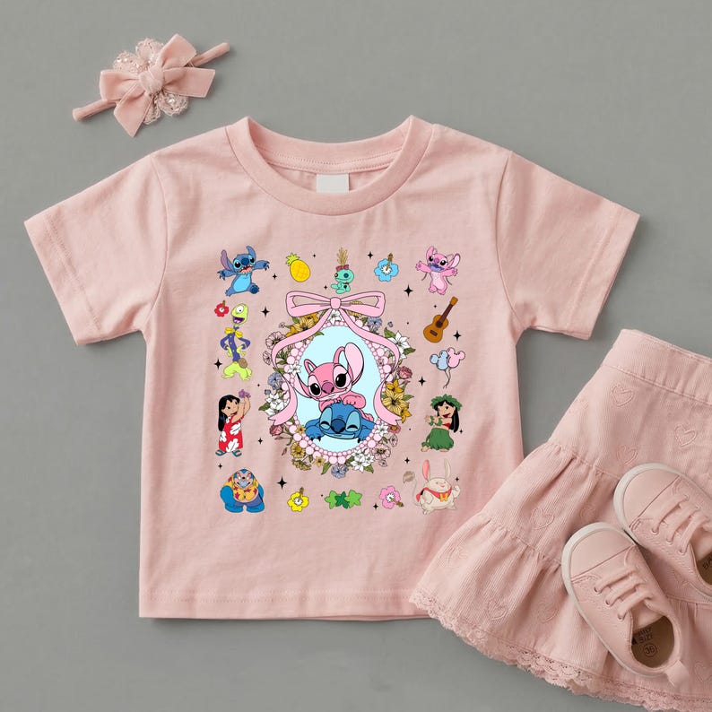 May include: A light pink short-sleeved t-shirt featuring a central oval frame with a pink bow, containing two blue and pink alien characters. The shirt is decorated with various small cartoon figures, flowers, a guitar, and a pineapple graphic.
