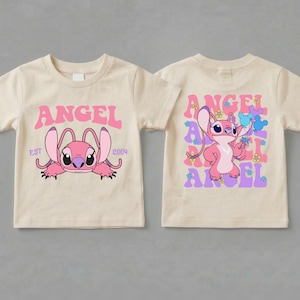 May include: A beige t-shirt featuring a pink Angel character graphic. The front displays the character with the text ANGEL EST 2004. The back shows the character holding blue balloons surrounded by the word ANGEL and small colourful flower icons.