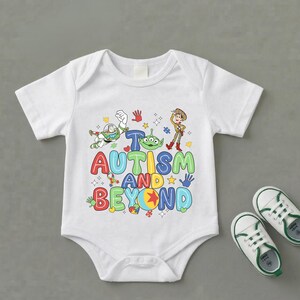 Op de afbeelding: Witte babybody met een kleurrijke afbeelding met personages en de tekst "Autisme en verder". Het ontwerp bevat Buzz Lightyear, Woody en een alien. Een paar witte en groene babyschoenen liggen naast de body.