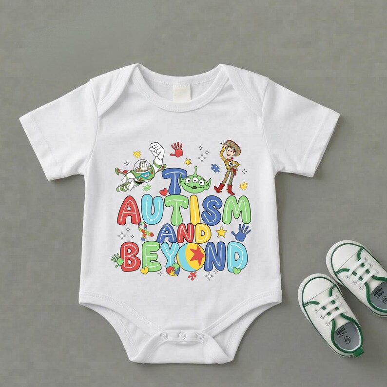 Op de afbeelding: Witte babybody met een kleurrijke afbeelding met personages en de tekst "Autisme en verder". Het ontwerp bevat Buzz Lightyear, Woody en een alien. Een paar witte en groene babyschoenen liggen naast de body.