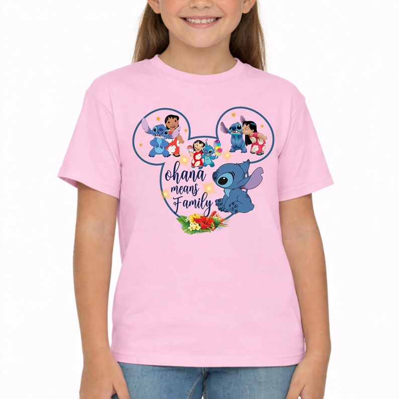 May include: A light pink short-sleeve t-shirt featuring a graphic of Stitch and Lilo inside a Mickey Mouse ear outline. The design includes the text "ohana means Family" in dark blue script, surrounded by small yellow stars and tropical flowers.