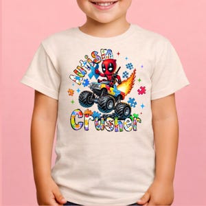 Puede incluir: Una camiseta beige claro con un gr&aacute;fico colorido de Deadpool conduciendo un monster truck con piezas de rompecabezas y las palabras "Autism Crusher". El dise&ntilde;o incluye llamas y estrellas.