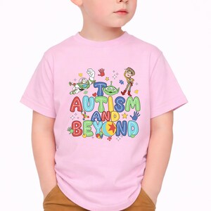 Op de afbeelding: Een lichtroze t-shirt met de woorden "To Autism and Beyond" in kleurrijke letters. Het shirt heeft stripfiguren en handafdrukken. Een leuk en ondersteunend ontwerp voor kinderen.