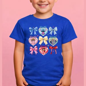 May include: A royal blue short-sleeved t-shirt featuring a central graphic print. The design displays a grid of various bows and heart-shaped frames containing cartoon characters, including Stitch, Angel, and Lilo, accented with small stars.