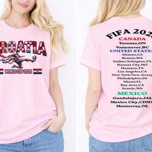 May include: Pink t-shirt featuring the word "CROATIA" in a red and white checkered design, with a soccer player graphic and "WORLD CUP 2026" text. The back of the shirt lists FIFA 2026 host cities in Canada, the United States, and Mexico.