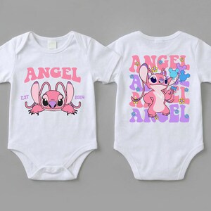 May include: Two white short-sleeved baby bodysuits. The left one features a pink Angel character with the text ANGEL EST 2004. The right one displays a standing pink Angel character surrounded by repeating purple ANGEL text and small colourful flowers.