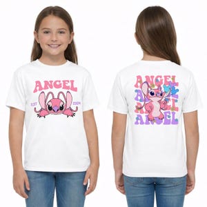 May include: A white t-shirt featuring a pink cartoon character on the front with the text ANGEL EST 2004. The back displays the same pink character surrounded by repeating purple and pink ANGEL text and small colourful flower graphics.