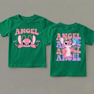 May include: A green short-sleeved t-shirt featuring a pink character graphic. The front displays the text ANGEL EST 2004. The back shows the character Angel surrounded by repeating ANGEL text, small flowers, and blue mouse-ear silhouettes.