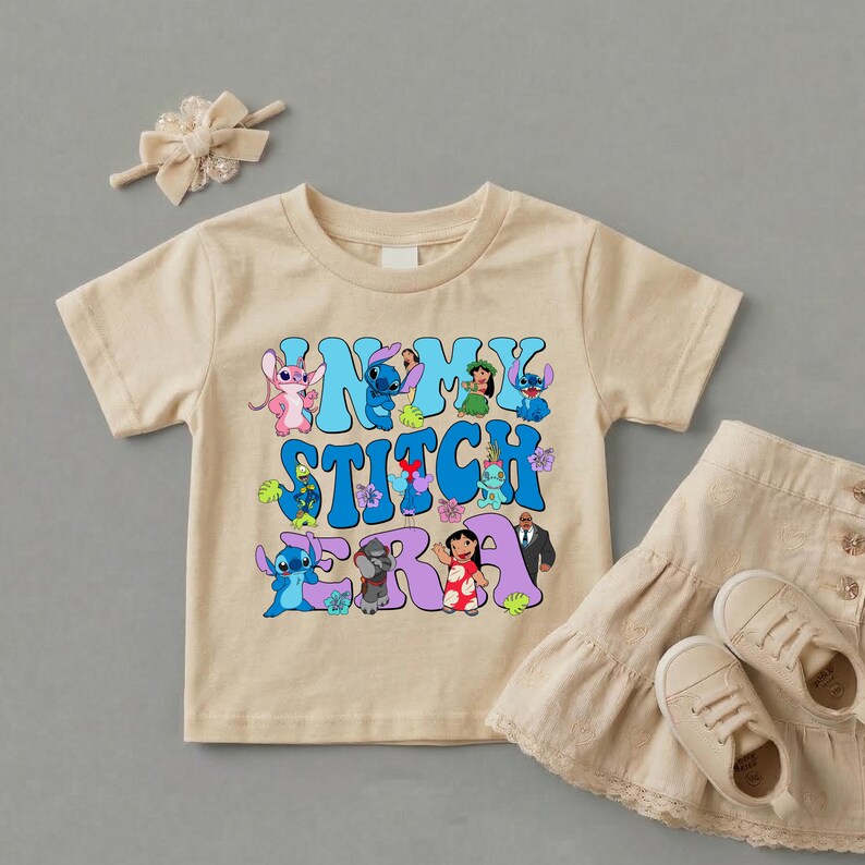 Pode incluir: Uma t-shirt bege de manga curta com uma estampa gr&aacute;fica com o texto "IN MY STITCH ERA" em letras azuis e roxas, rodeado por v&aacute;rias personagens de Lilo e Stitch, acompanhada por um la&ccedil;o de cabelo e uma saia bege com folhos e t&eacute;nis.