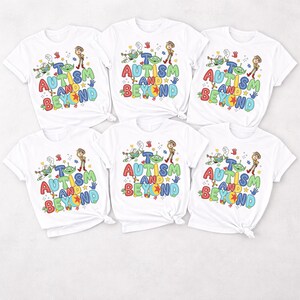Op de afbeelding: Zes witte t-shirts met een kleurrijk grafisch ontwerp. Het ontwerp bevat de tekst "AUTISM AND BEYOND" in verschillende kleuren, samen met stripfiguren. De shirts zijn korte mouwen en hebben een ronde hals.