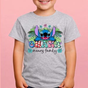 May include: A light gray t-shirt featuring a graphic of the blue character Stitch surrounded by green tropical leaves and hibiscus flowers. The text OHANA is printed in colorful patterned letters, with the words means family below in black script.