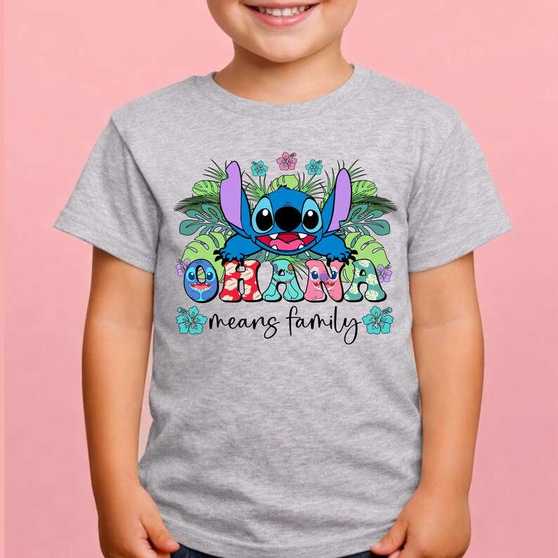 May include: A light gray t-shirt featuring a graphic of the blue character Stitch surrounded by green tropical leaves and hibiscus flowers. The text OHANA is printed in colorful patterned letters, with the words means family below in black script.