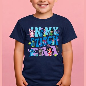 Pode incluir: Uma t-shirt azul-marinho de manga curta com uma estampa gr&aacute;fica que inclui o texto "IN MY STITCH ERA" em azul-claro e roxo. O design apresenta v&aacute;rias personagens de desenhos animados da franquia Lilo e Stitch e flores tropicais.