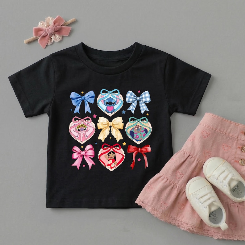 May include: A black short-sleeved t-shirt featuring a graphic print of various bows and heart-shaped frames containing cartoon characters. A pink hair bow with a pearl accent and a pink skirt with white sneakers are also visible in the image.