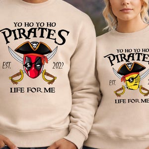 K&ouml;nnte beinhalten: Beige Sweatshirts mit Piratenmotiv. Das Design zeigt einen Piratenhut, gekreuzte Schwerter und den Text "YO HO YO HO PIRATES EST. 2022 LIFE FOR ME." Ein Sweatshirt hat einen von Deadpool inspirierten Piraten, das andere einen gelben Piraten.