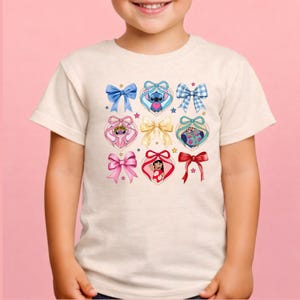 May include: A cream-colored short-sleeve t-shirt featuring a graphic print of various bows and heart-shaped frames. Inside the hearts are illustrations of Stitch, Angel, Lilo, and Scrump, accented by small stars and a central gold-colored bow.