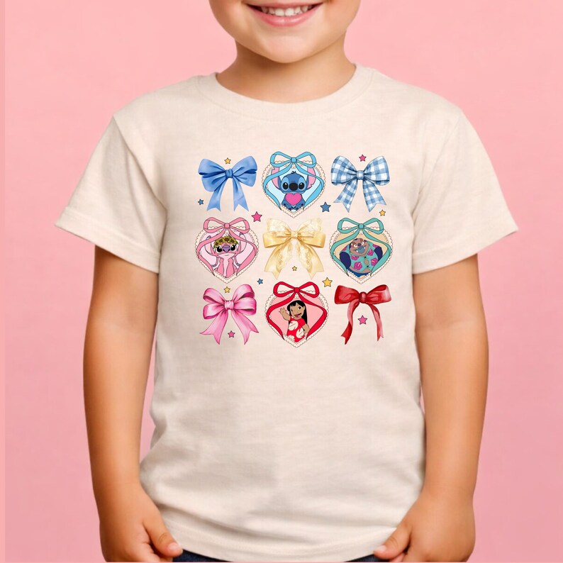 May include: A cream-colored short-sleeve t-shirt featuring a graphic print of various bows and heart-shaped frames. Inside the hearts are illustrations of Stitch, Angel, Lilo, and Scrump, accented by small stars and a central gold-colored bow.
