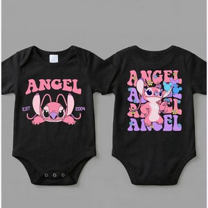 May include: Two black baby bodysuits featuring a pink character graphic. The left bodysuit displays the text ANGEL EST 2004. The right bodysuit shows the character Angel surrounded by the word ANGEL in a repeating pattern with small colourful flowers.