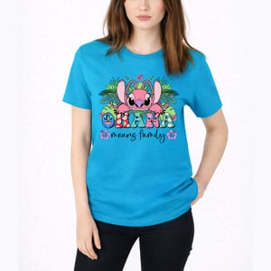 May include: A bright turquoise short-sleeve t-shirt featuring a graphic of a pink alien character surrounded by tropical leaves and flowers. The text "OHANA" is printed in colorful patterned letters above the script text "means family".