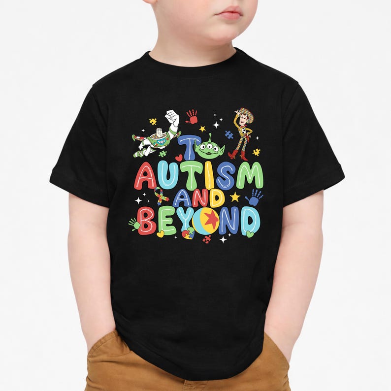 Op de afbeelding: Zwart T-shirt met een kleurrijke afbeelding met de tekst "Autisme en verder". Het ontwerp bevat stripfiguren en symbolen. Het shirt wordt door een kind gedragen.