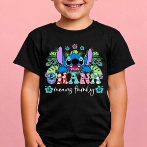 May include: A black t-shirt featuring a graphic of the blue character Stitch surrounded by green tropical leaves and colorful hibiscus flowers. The text OHANA is printed in patterned letters, with the phrase means family written below in white.