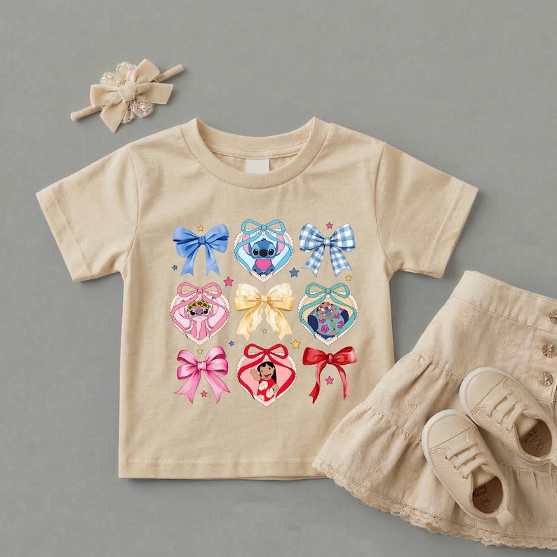 May include: A beige short-sleeve t-shirt featuring a graphic print of various bows and heart-shaped frames containing cartoon characters. The set includes a matching beige hair bow and a beige ruffled skirt with two beige sneakers.